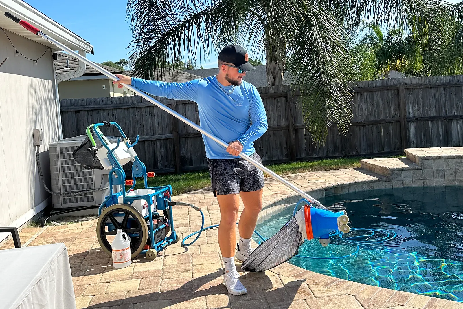 Pool tech vacuuming a backyard pool with a service cart on a weekly visit